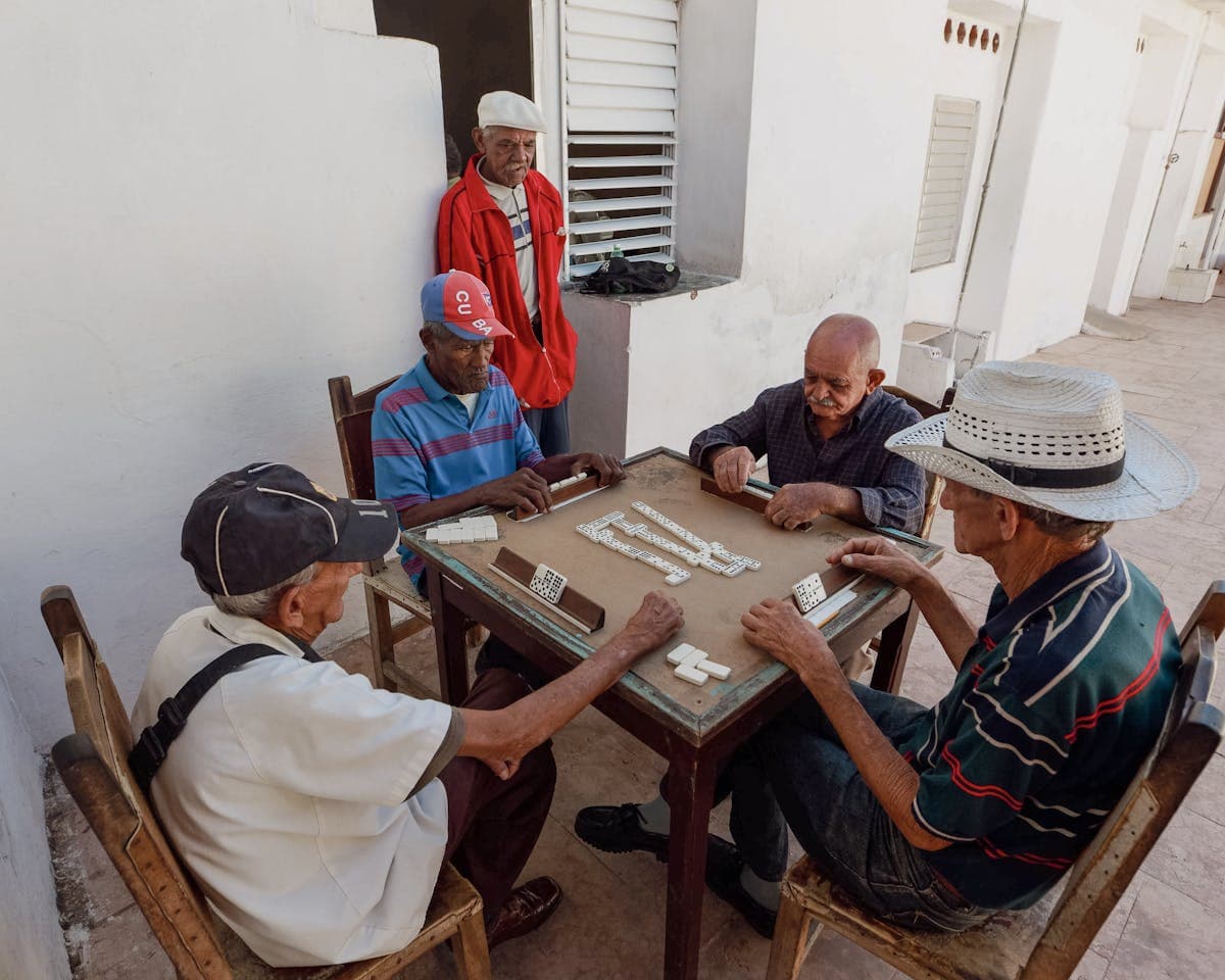 Four men playing dominoes at a table outside a colmado, whitewashed walls in the background